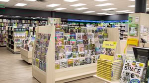 Interior of a Paper Plus store showing shelves of magazines, books, and stationery under bright lighting.