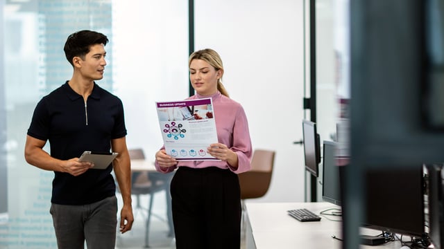 A man and woman walking through an office while reviewing an A3 colour printed business update document and a tablet.