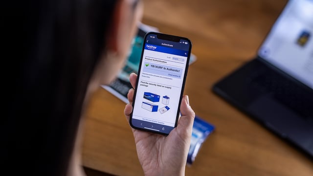 A person views a Brother product authentication page on a smartphone showing a confirmation that the consumable is authentic, with printed materials and a laptop on the desk.