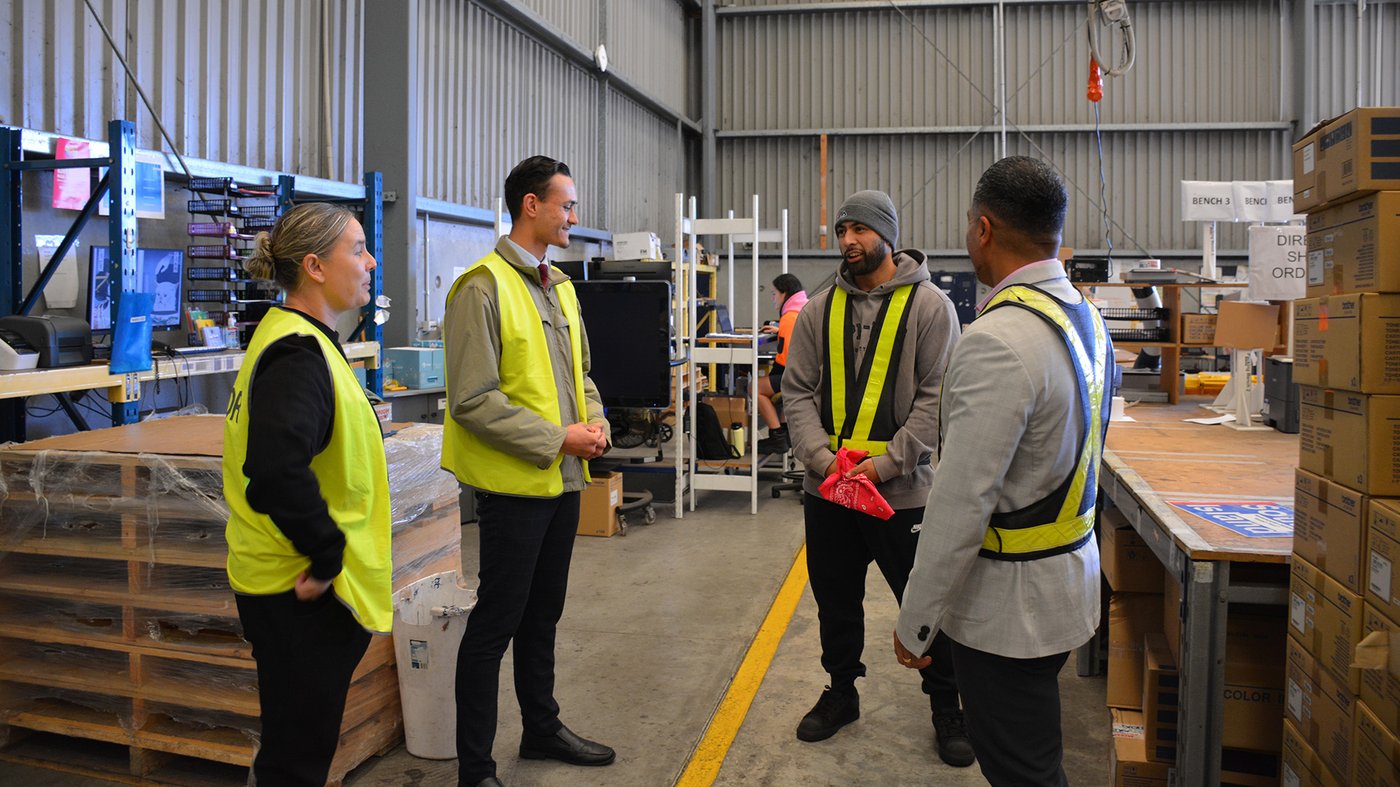 Group of staff and a youth worker wearing high visibility vests talking together inside a warehouse as part of a community development program.