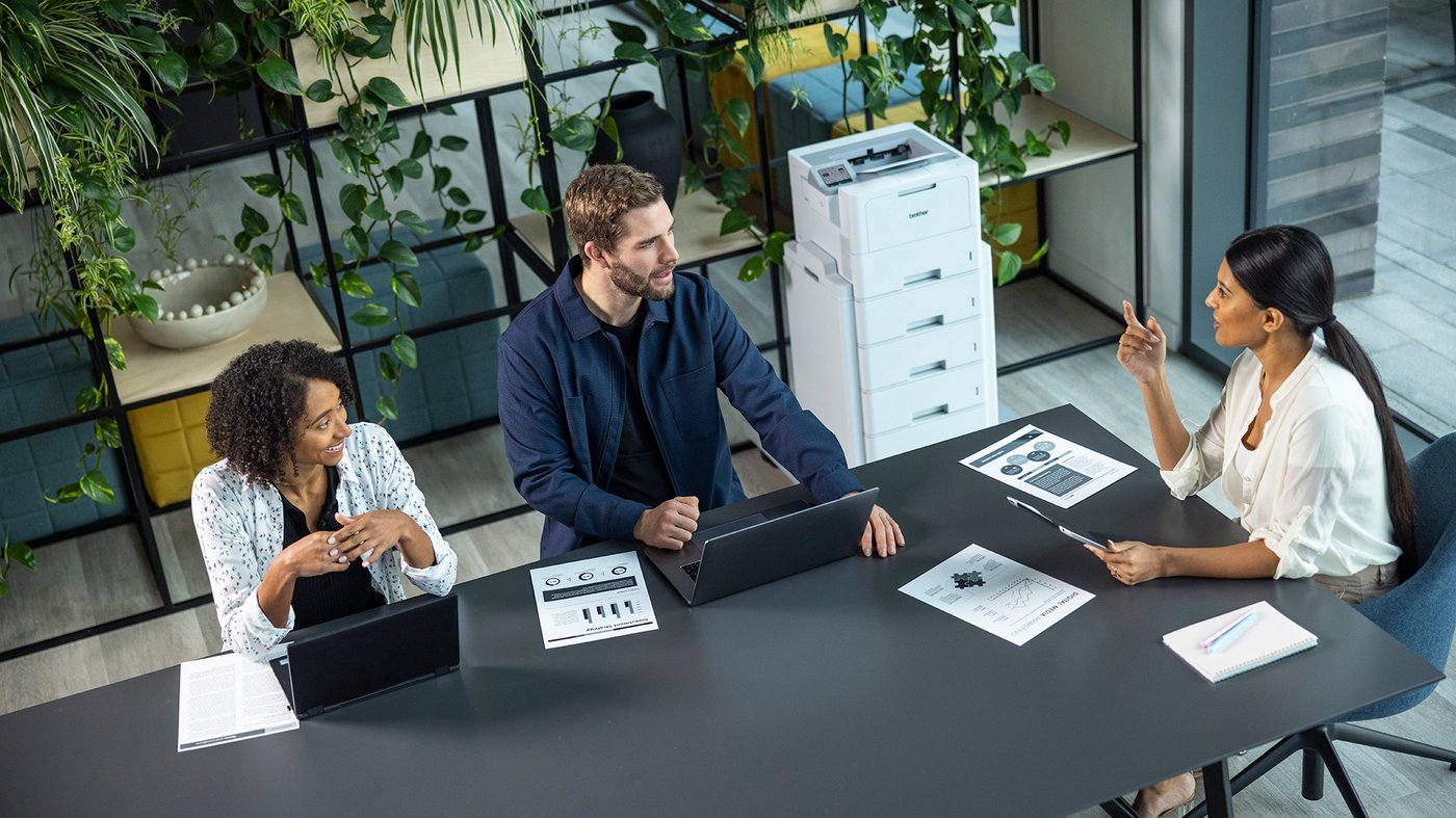 Three colleagues sit around a meeting table with laptops and printed documents, discussing work in a modern office with a Brother printer in the background.