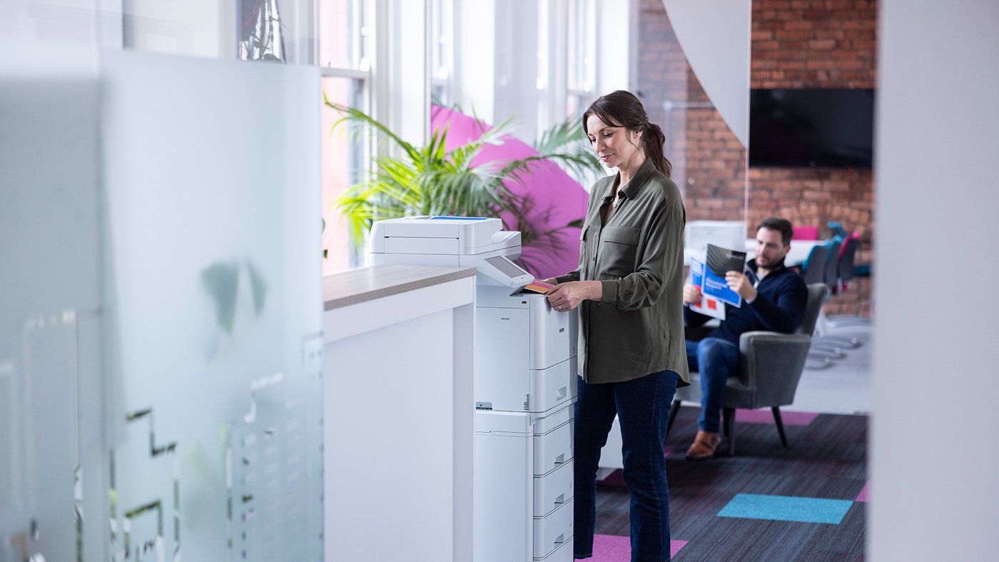 A woman uses a Brother multifunction printer at an office counter while a man reads printed materials in the seating area behind her.