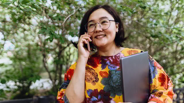 Smiling woman salesperson outdoors holding a laptop and talking on a mobile phone, standing in front of leafy trees.