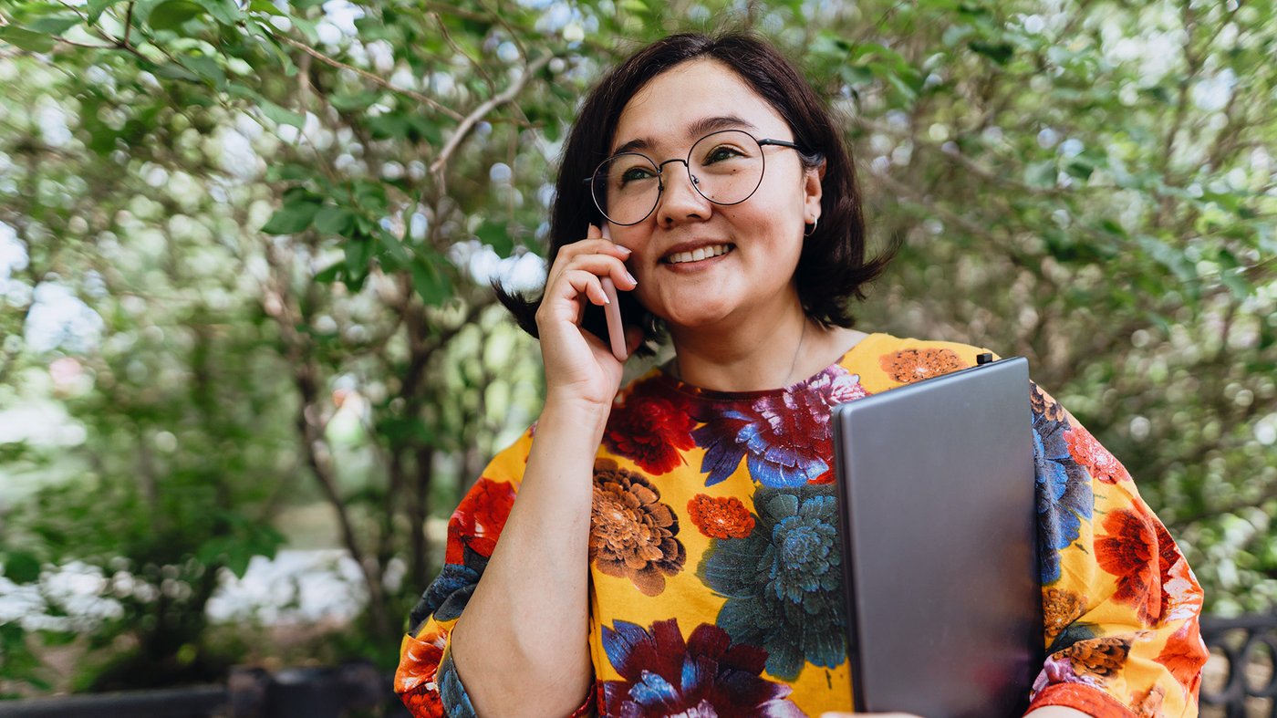 Smiling woman salesperson outdoors holding a laptop and talking on a mobile phone, standing in front of leafy trees.