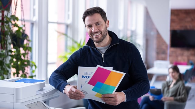 A man standing beside a printer holds several colour printouts and smiles at the camera.