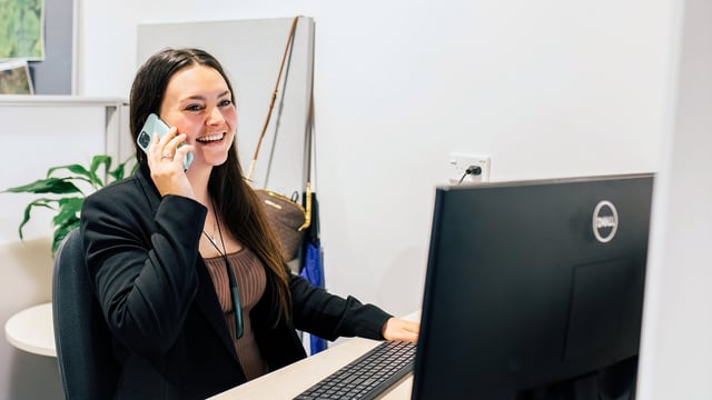 A woman sitting at a desk smiles while speaking on the phone, with a computer monitor and office items around her.