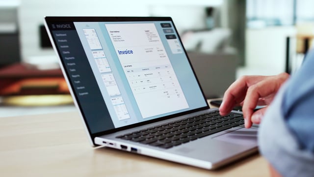 A person works on a laptop displaying invoice creation software, with an invoice preview open on the screen in a modern office setting.