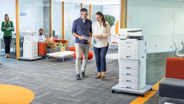 People work and move through a modern office space with two Brother multifunction printers positioned in common areas.