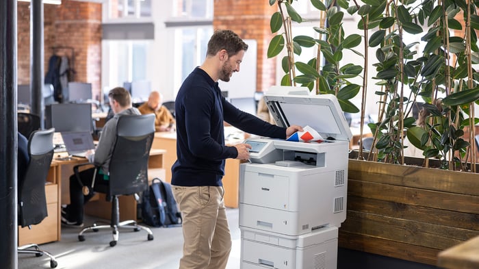 Man standing at a multifunction printer in a modern open plan office, placing documents on the scanner while colleagues work at nearby desks.
