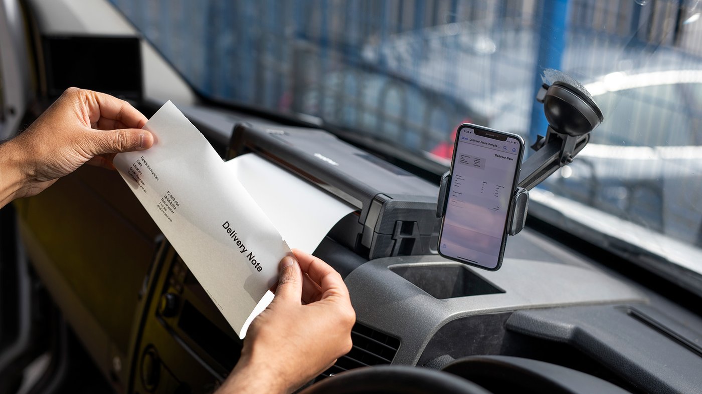 Person removing a printed delivery note from a Brother mobile printer mounted in a vehicle, with a smartphone displaying the document on the dashboard.