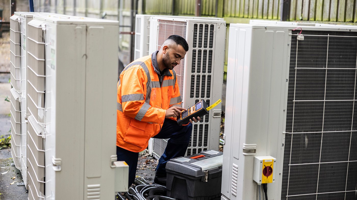Technician in a high visibility jacket using a handheld label printer while working on outdoor HVAC units.
