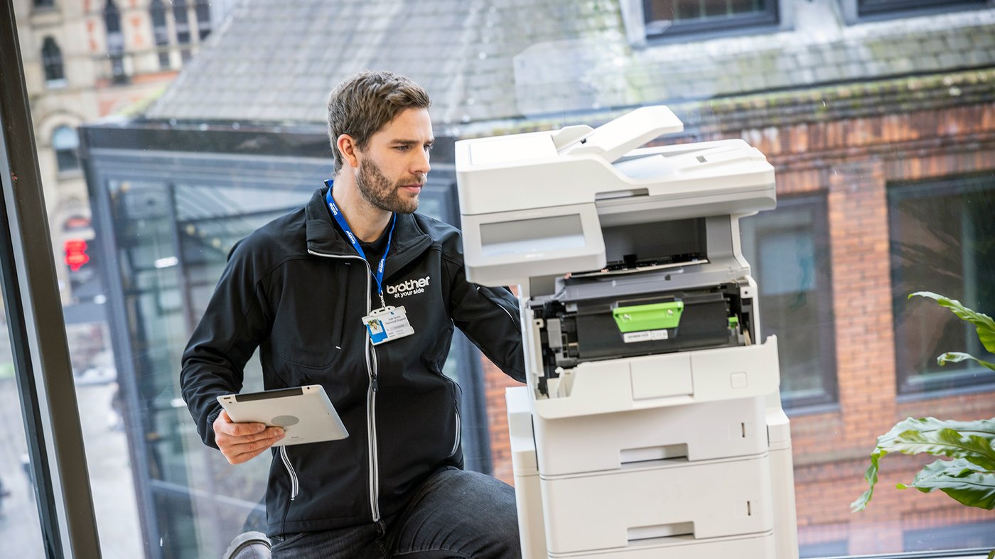 Brother service technician kneeling beside an open multifunction printer, inspecting the internal components while holding a tablet.