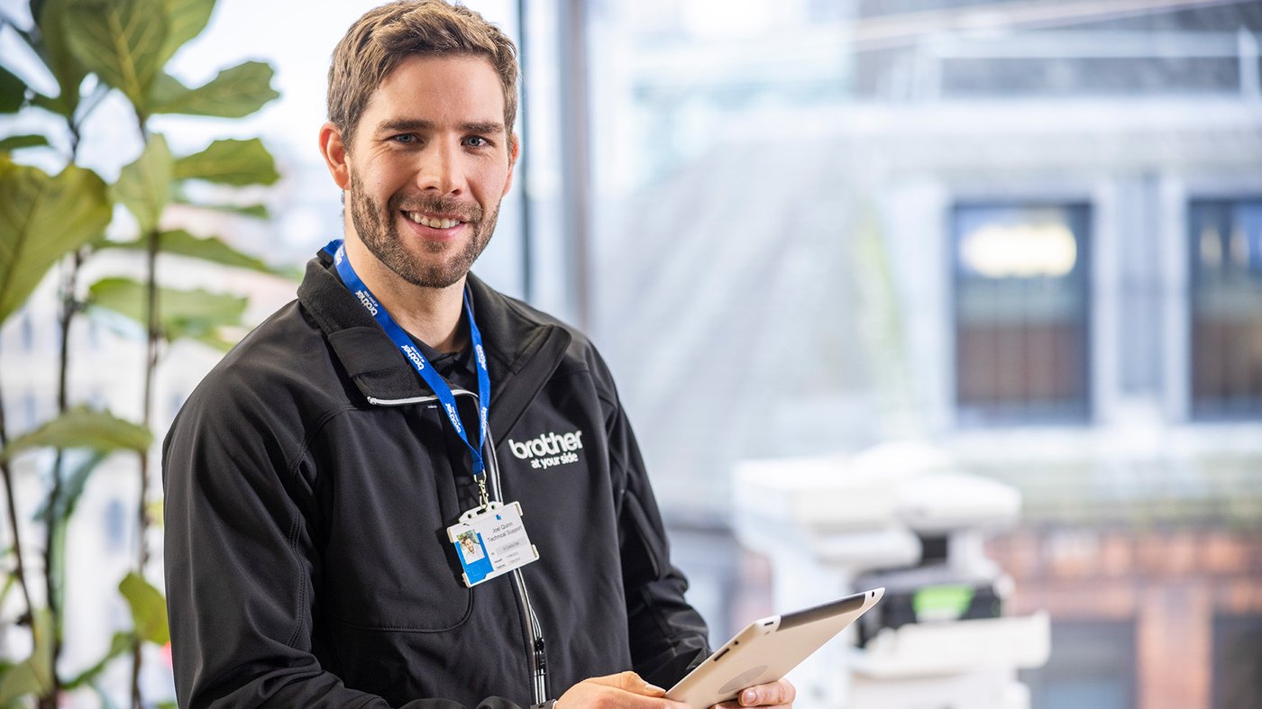 Brother service technician smiling at the camera while holding a tablet, with a printer and office window behind him.