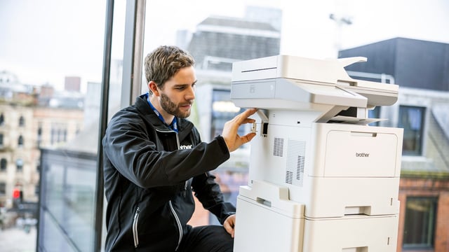 Brother service technician adjusting a multifunction printer inside an office with large windows and city buildings in the background.