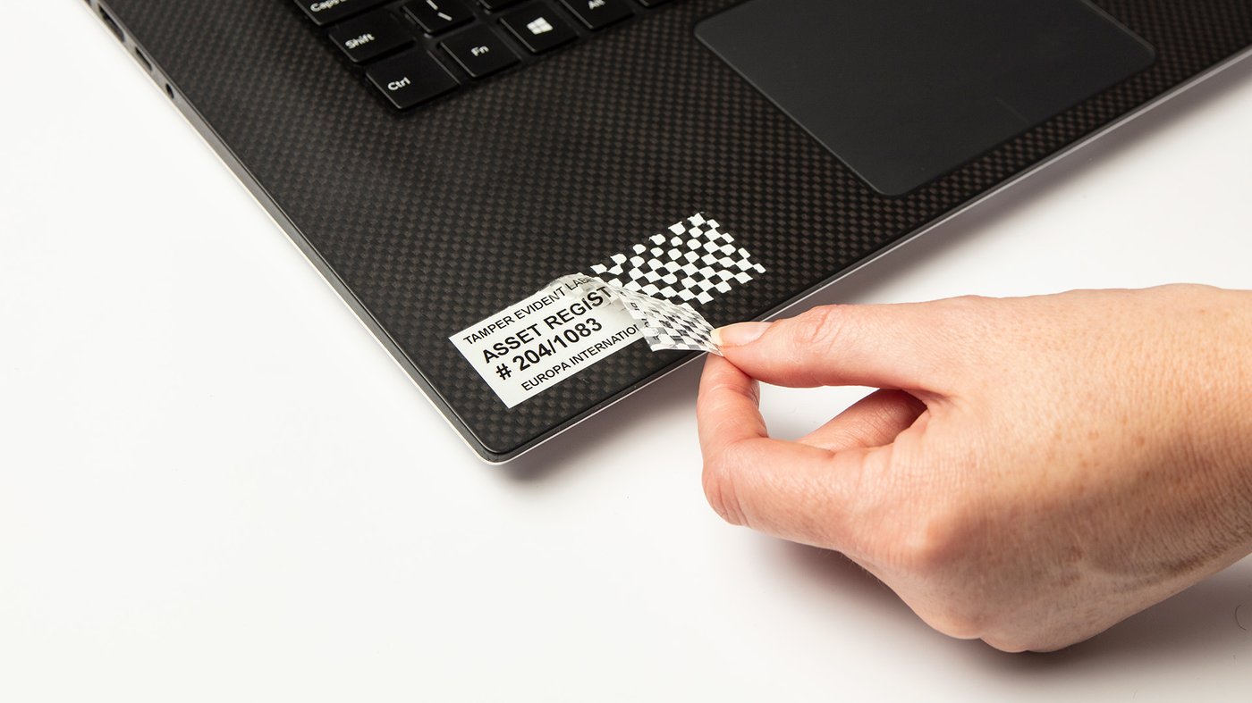 Close up of a hand peeling a tamper evident security label from a laptop, revealing a checkerboard pattern indicating tampering.