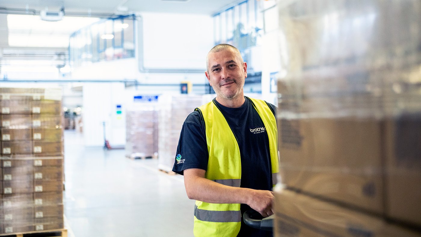 Warehouse worker wearing a high visibility vest moving packaged goods in a Brother supply chain facility.