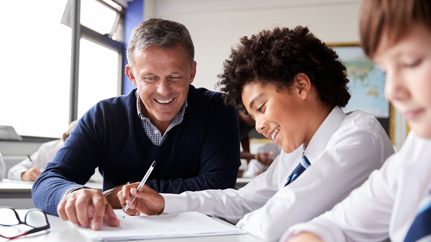 A teacher sits beside a student in uniform, smiling as they work together on an assignment in class.