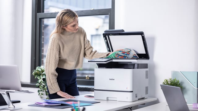 Woman placing a document on the scanner of a Brother colour laser printer in an office workspace.