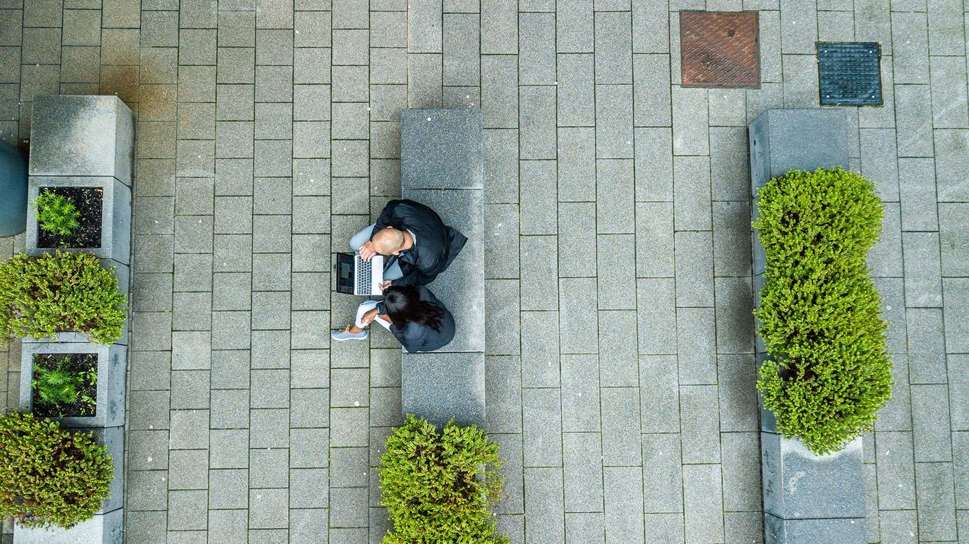 Overhead view of two people sitting on a stone bench outdoors, working together on a laptop between rows of neatly trimmed bushes.