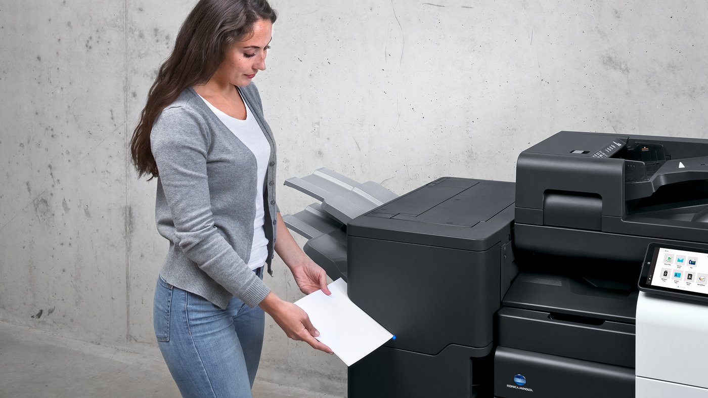 A woman collects printed pages from a Konica Minolta bizhub C651i multifunction printer in a modern office setting.