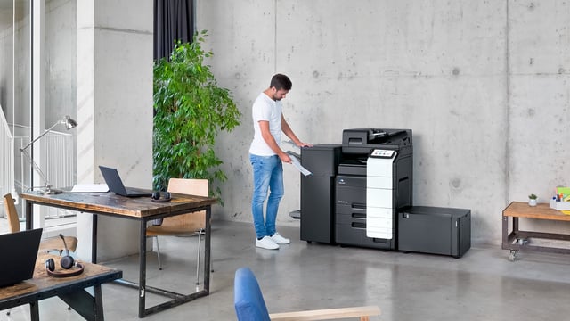 A man retrieves printed pages from a Konica Minolta bizhub C651i printer in a bright, modern workspace with industrial design features.