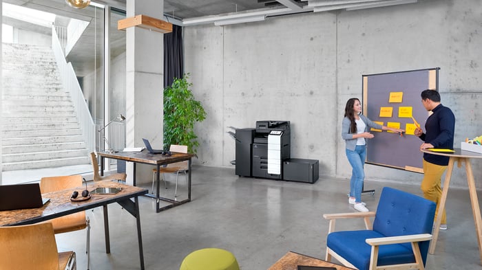Two colleagues collaborate at a board covered in sticky notes while a Konica Minolta bizhub C651i printer sits in the background of the open office.