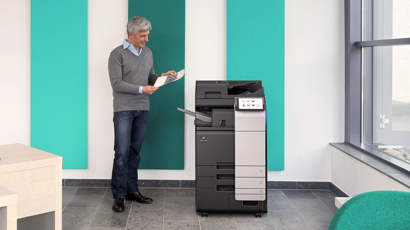 Person standing beside a Konica Minolta bizhub C301i multifunction printer reviewing freshly printed documents in a modern office.