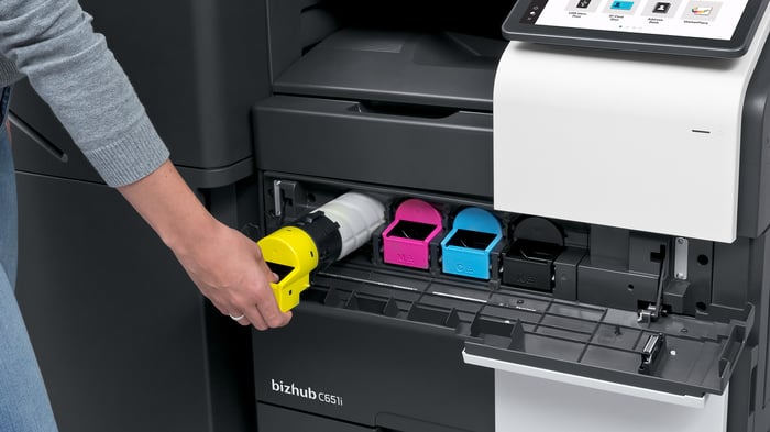 A person replaces the yellow toner cartridge inside a Konica Minolta bizhub C651i printer, with the magenta, cyan, and black toner slots visible.