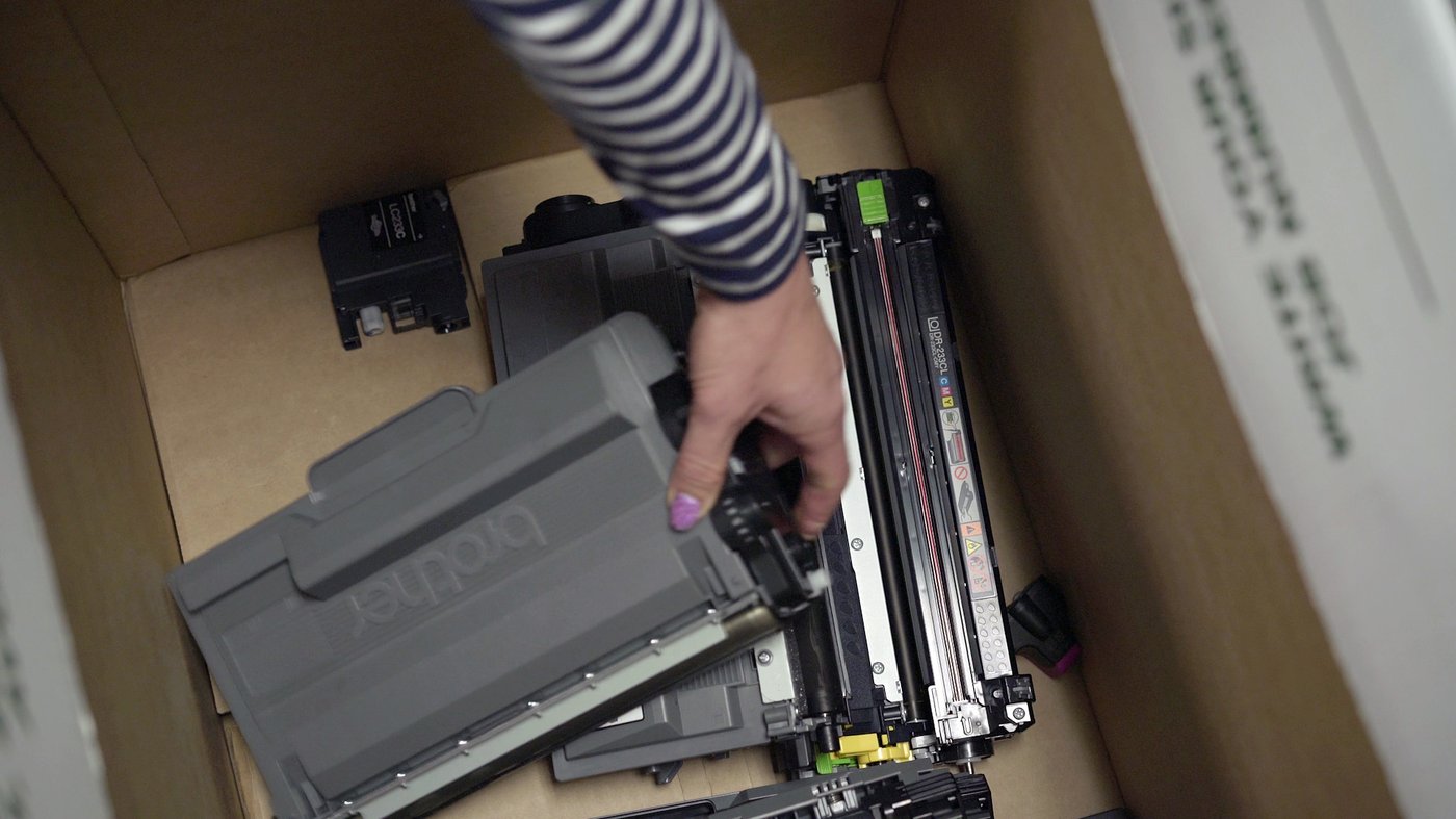 Hand placing used Brother printer cartridges into a cardboard recycling box.
