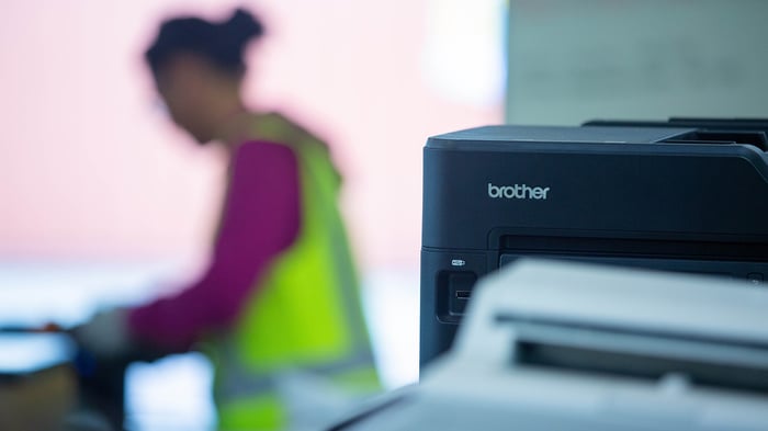 Close-up of a Brother printer in the foreground with a person in high-visibility workwear blurred in the background.