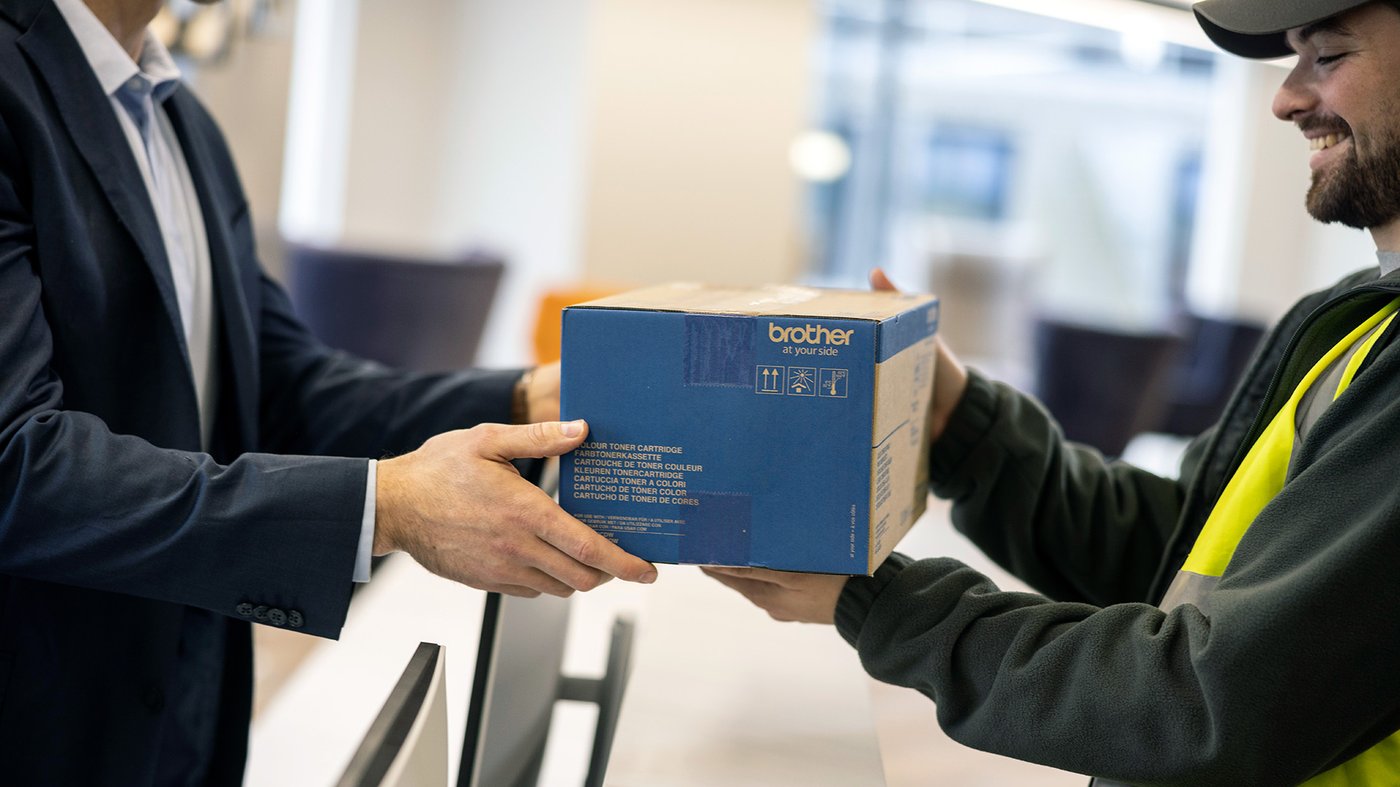 A delivery worker hands a Brother branded carton to an office employee across a counter.