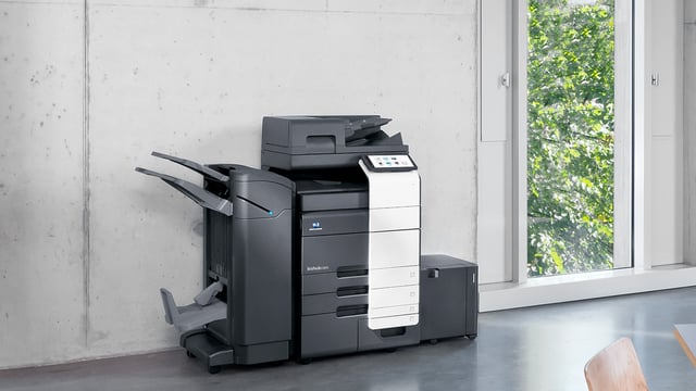 Konica Minolta bizhub office multifunction copier placed against a concrete wall beside a large window with greenery outside.