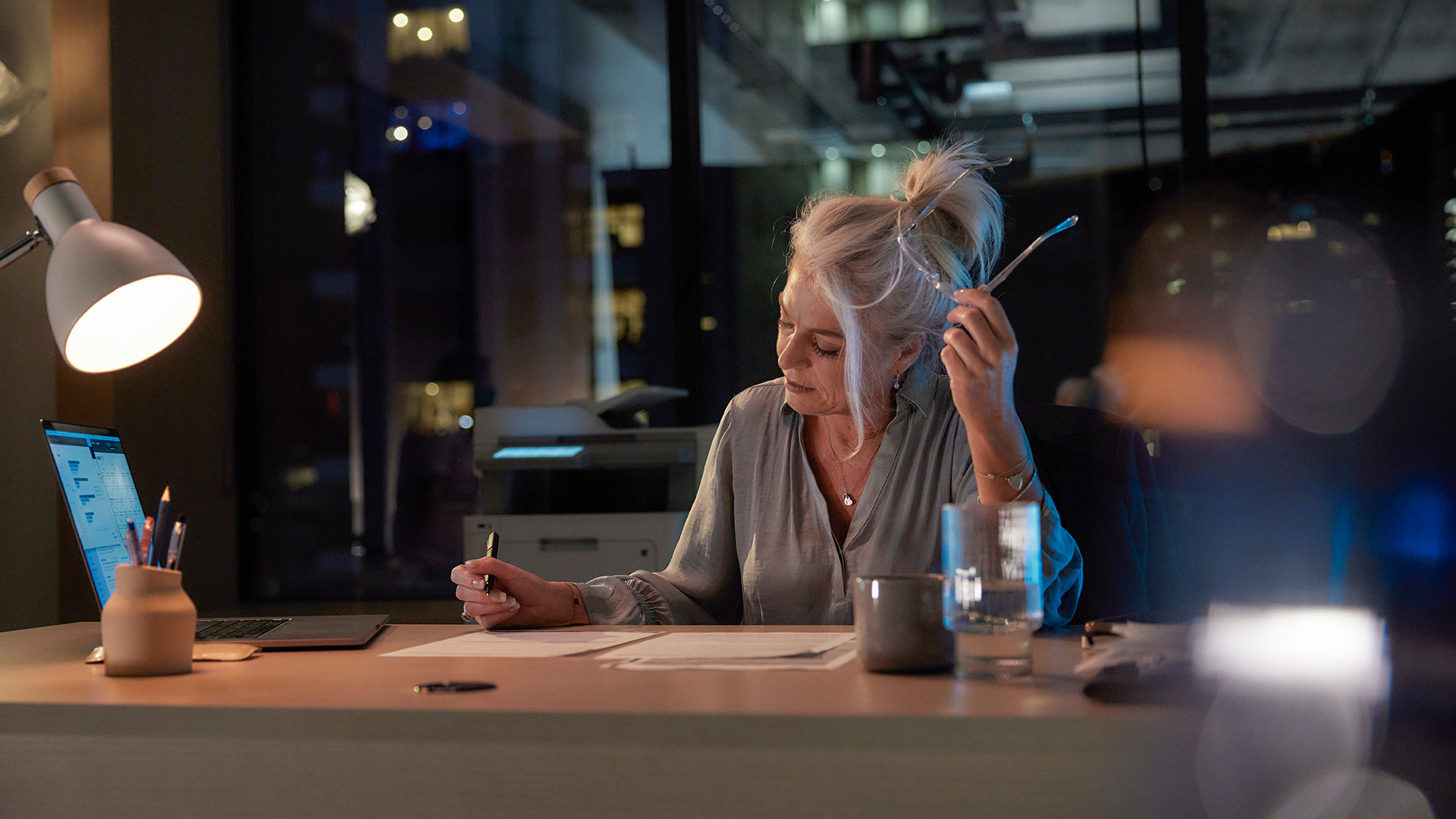 A woman with grey hair works late at her desk, reviewing documents under a desk lamp with a laptop open beside her.
