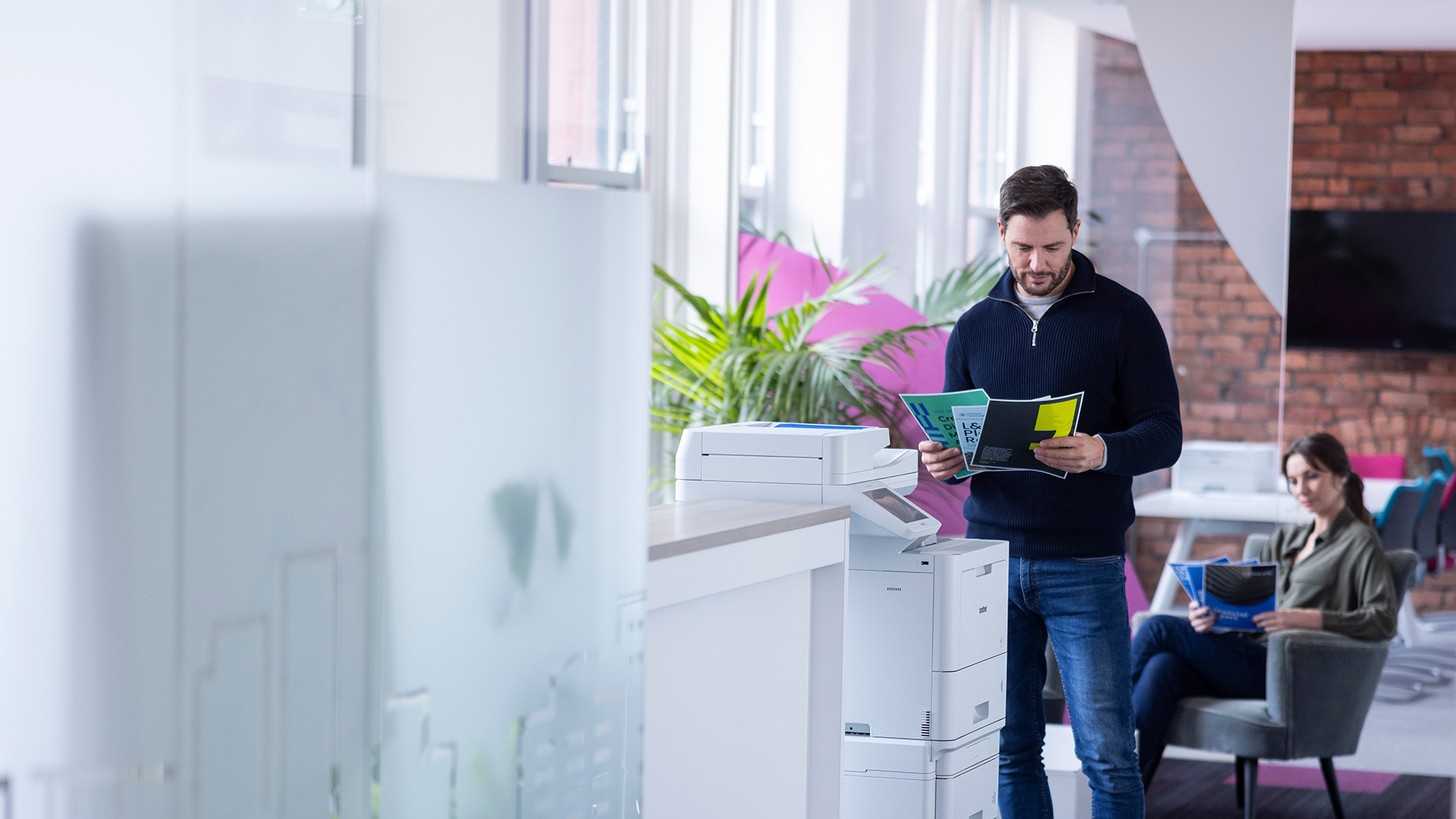 Man standing at a multifunction printer in a modern office while reading printed documents, with a woman seated in the background reviewing papers.