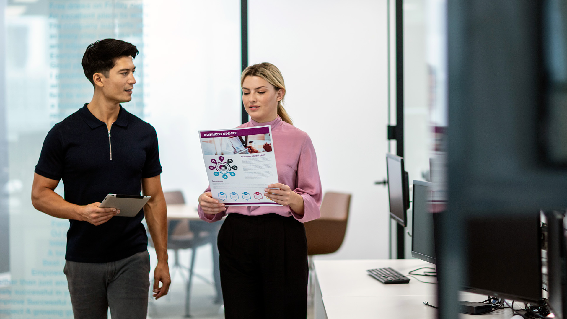 A man and woman walking through an office while reviewing an A3 colour printed business update document and a tablet.