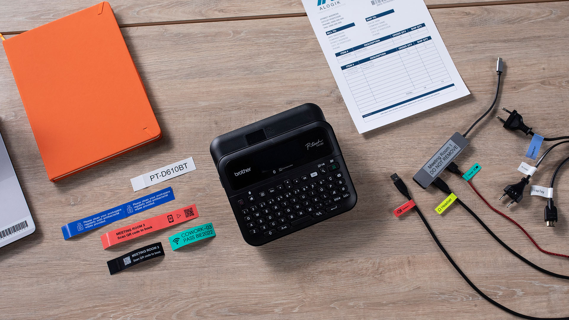 Desktop workspace with a Brother label maker, printed labels, laptop, notebook and cables organised with colour coded labels.