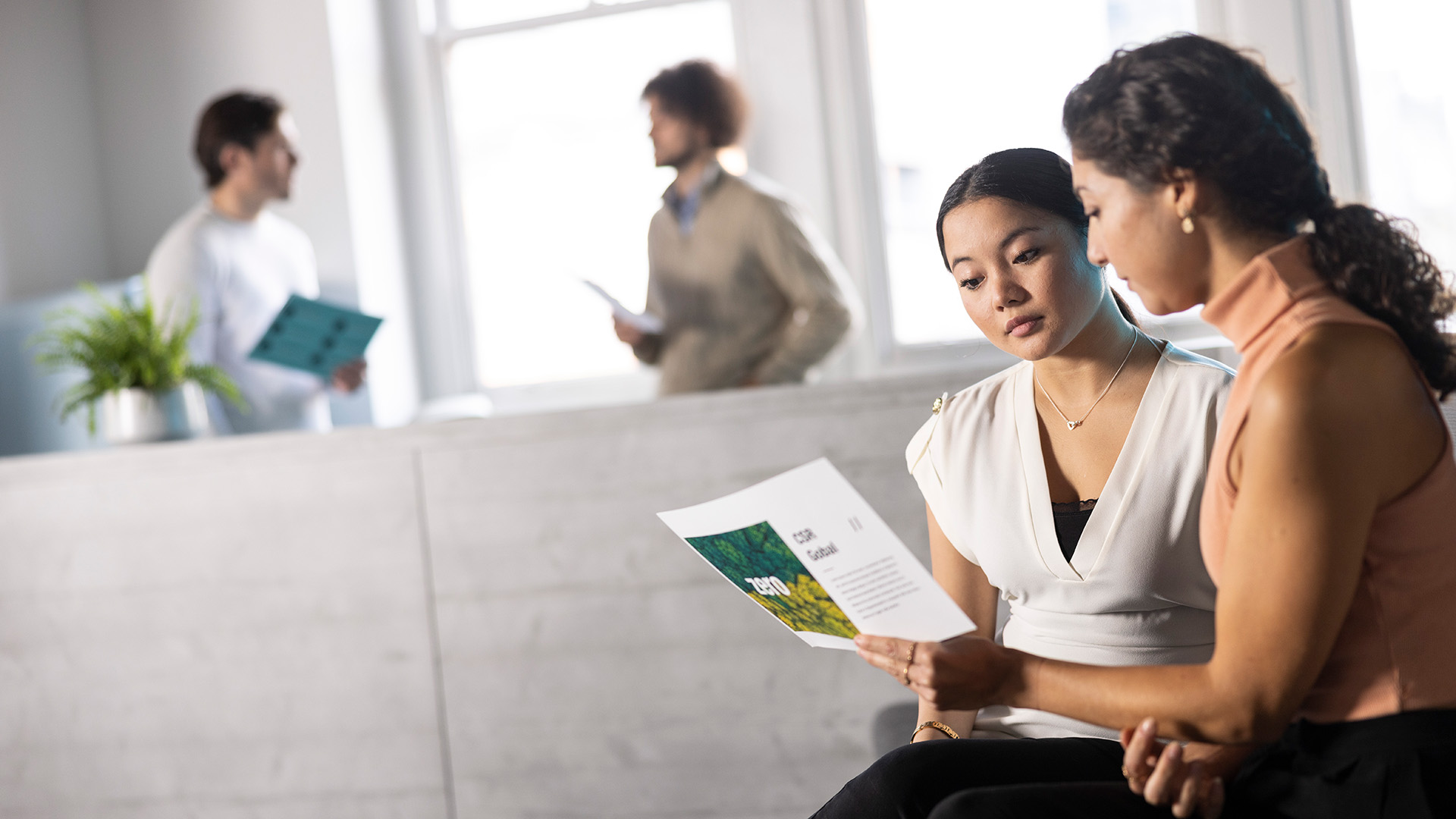 Two people seated together reviewing a printed document, with two others talking in the background of a bright office space.