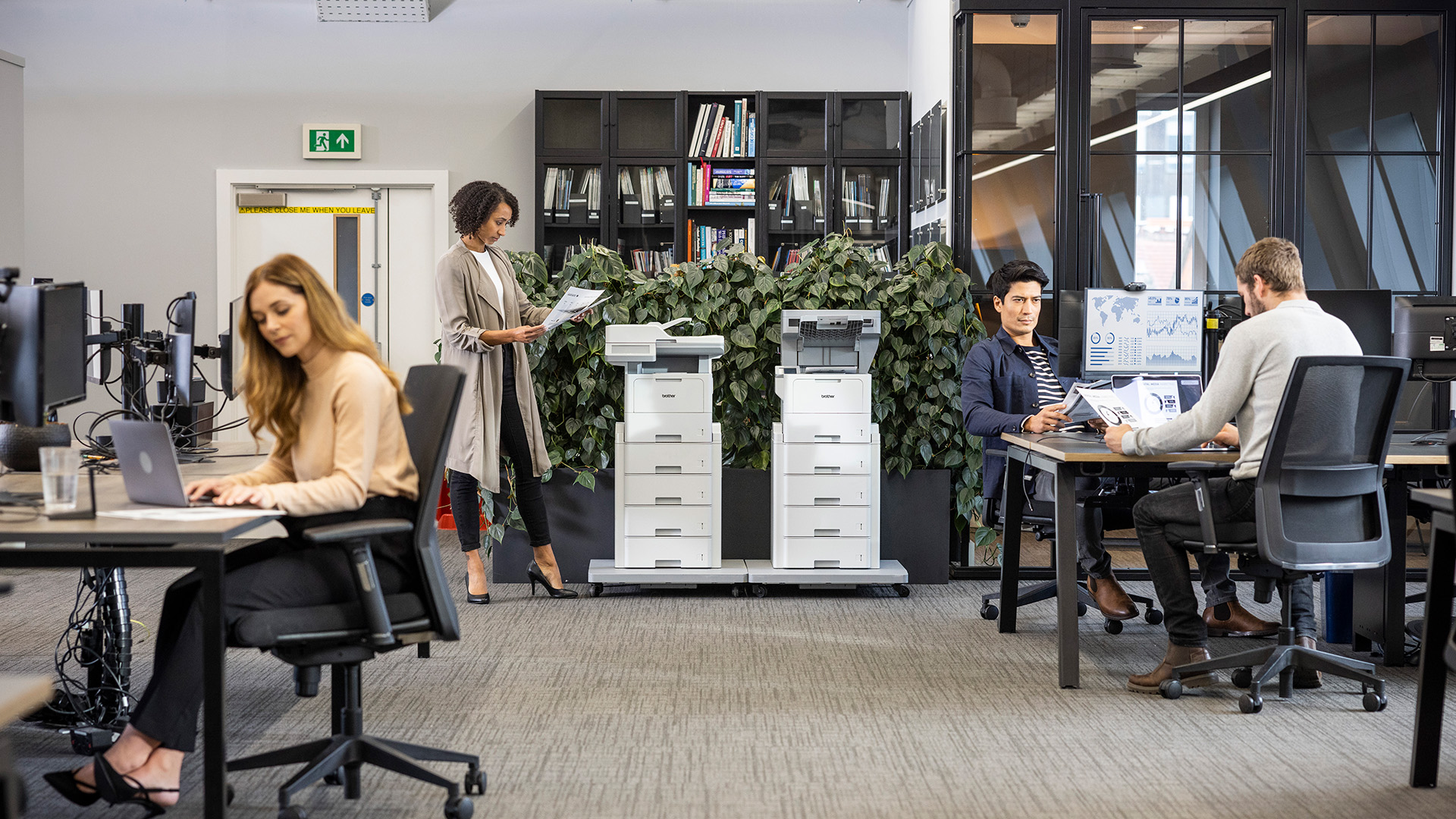 A busy office with people working at desks, while two Brother multifunction printers stand against a backdrop of indoor plants.