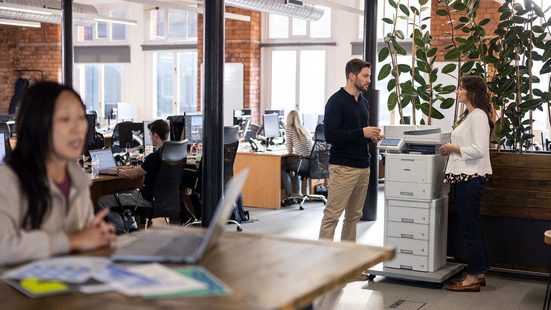 Two colleagues talking beside a Brother multifunction printer in a modern open-plan office, with other staff working at desks in the background.