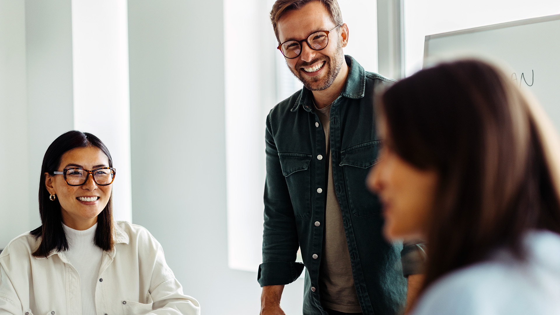 Two colleagues smiling and talking during a meeting, with a man standing and a woman seated at a table.