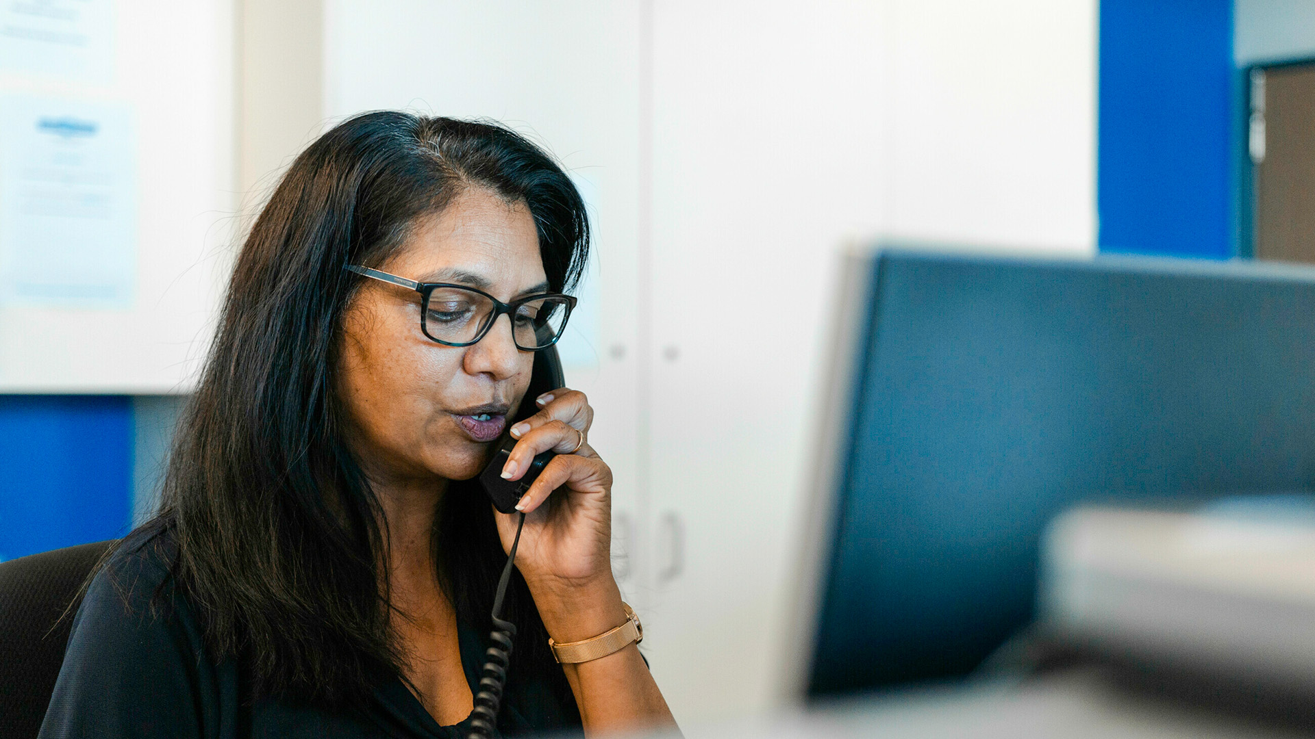 A woman wearing glasses speaks on a corded phone while sitting at a desk in an office environment, with a computer monitor in front of her.