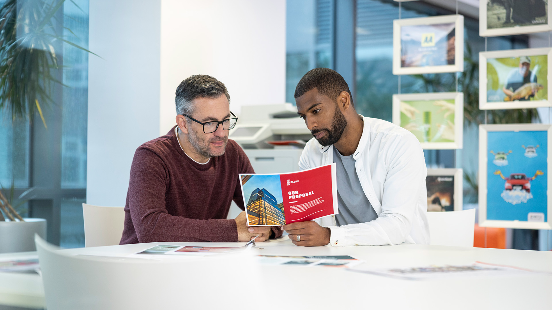 Two people sit together reviewing a printed proposal document in a bright, modern office.