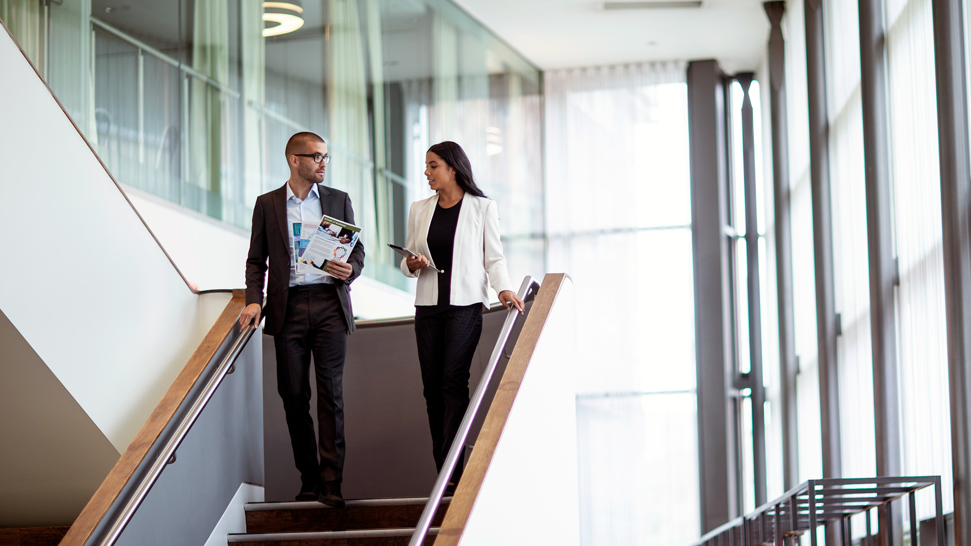 Two professionals walking down a staircase in a bright office building, talking while holding printed documents and a tablet.