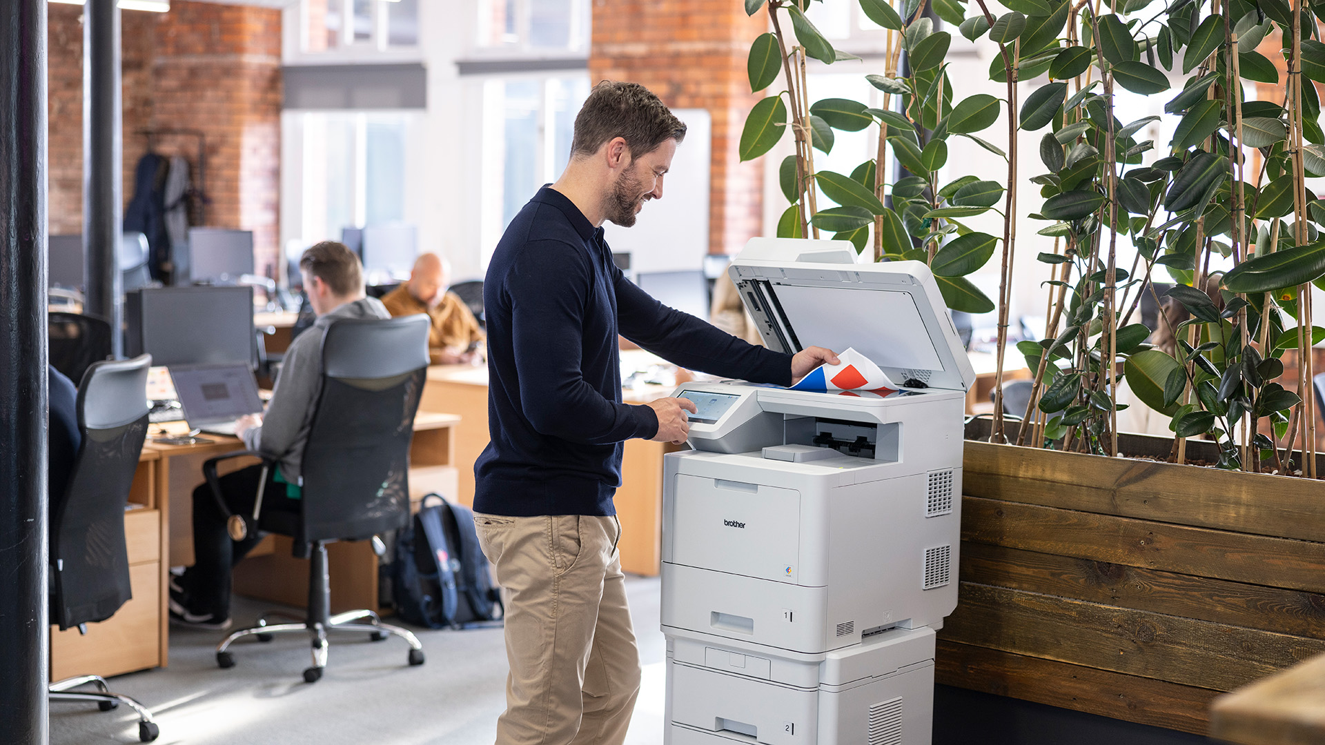 Man standing at a multifunction printer in a modern open plan office, placing documents on the scanner while colleagues work at nearby desks.
