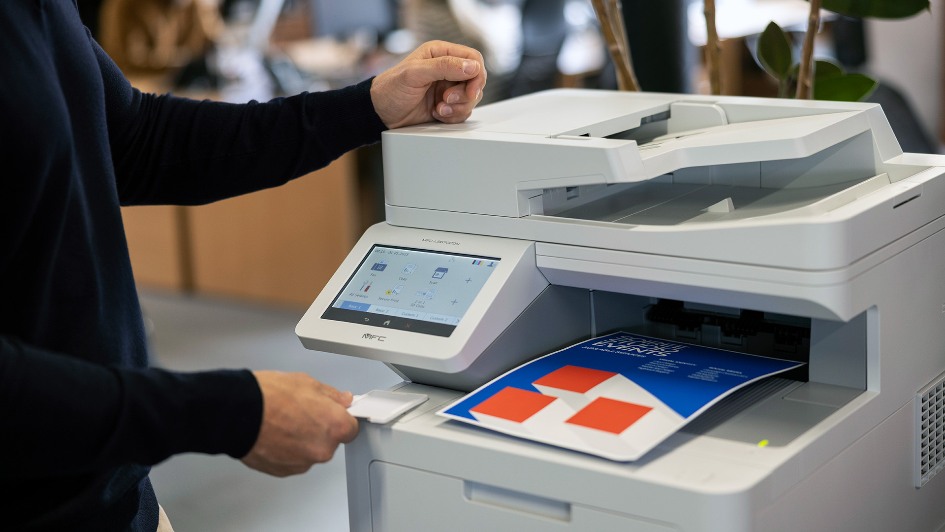 Person using a multifunction printer in an office, retrieving a colour document from the output tray.
