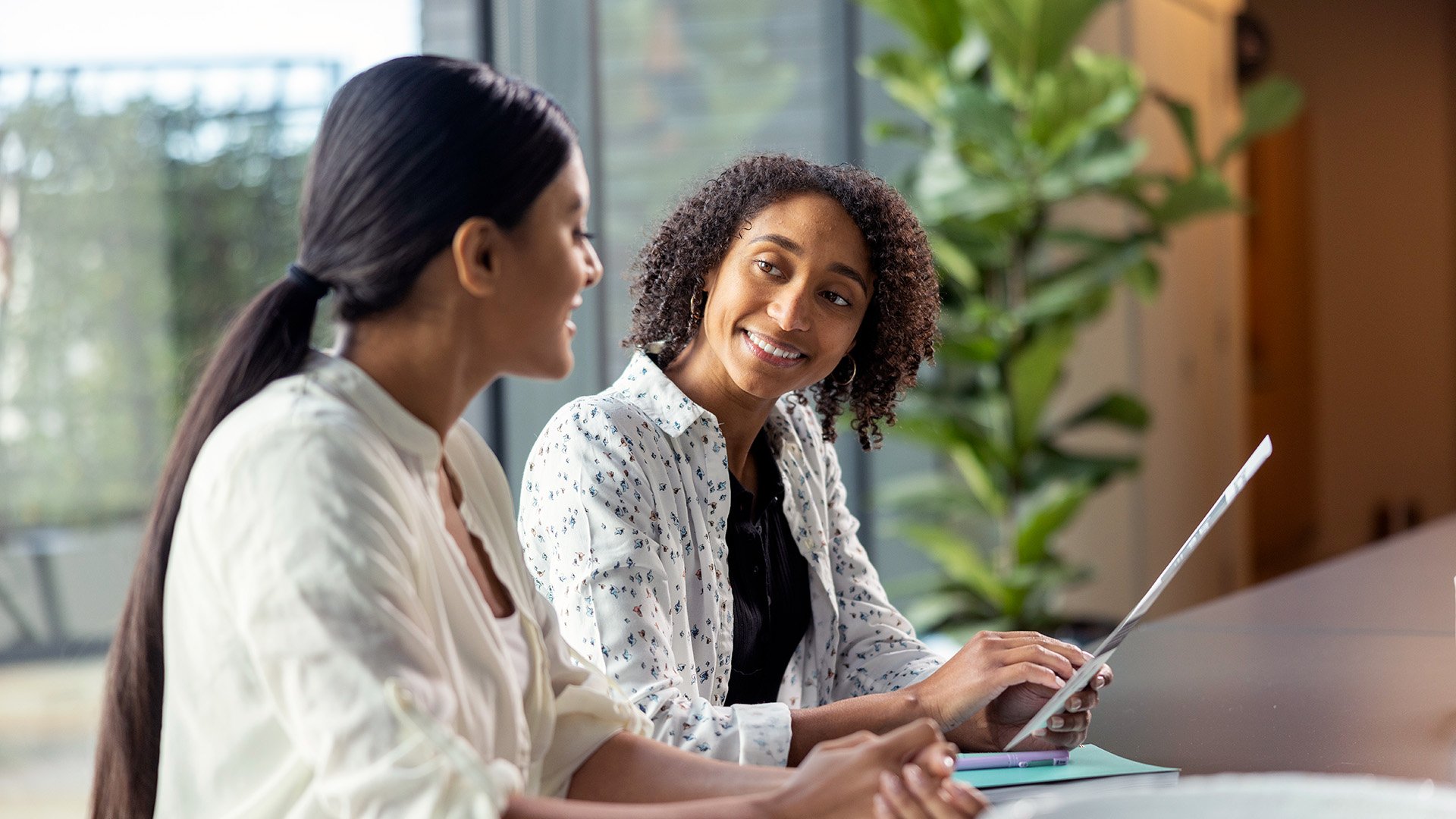 Two women sitting at a table in a modern office, smiling and talking while reviewing a document.