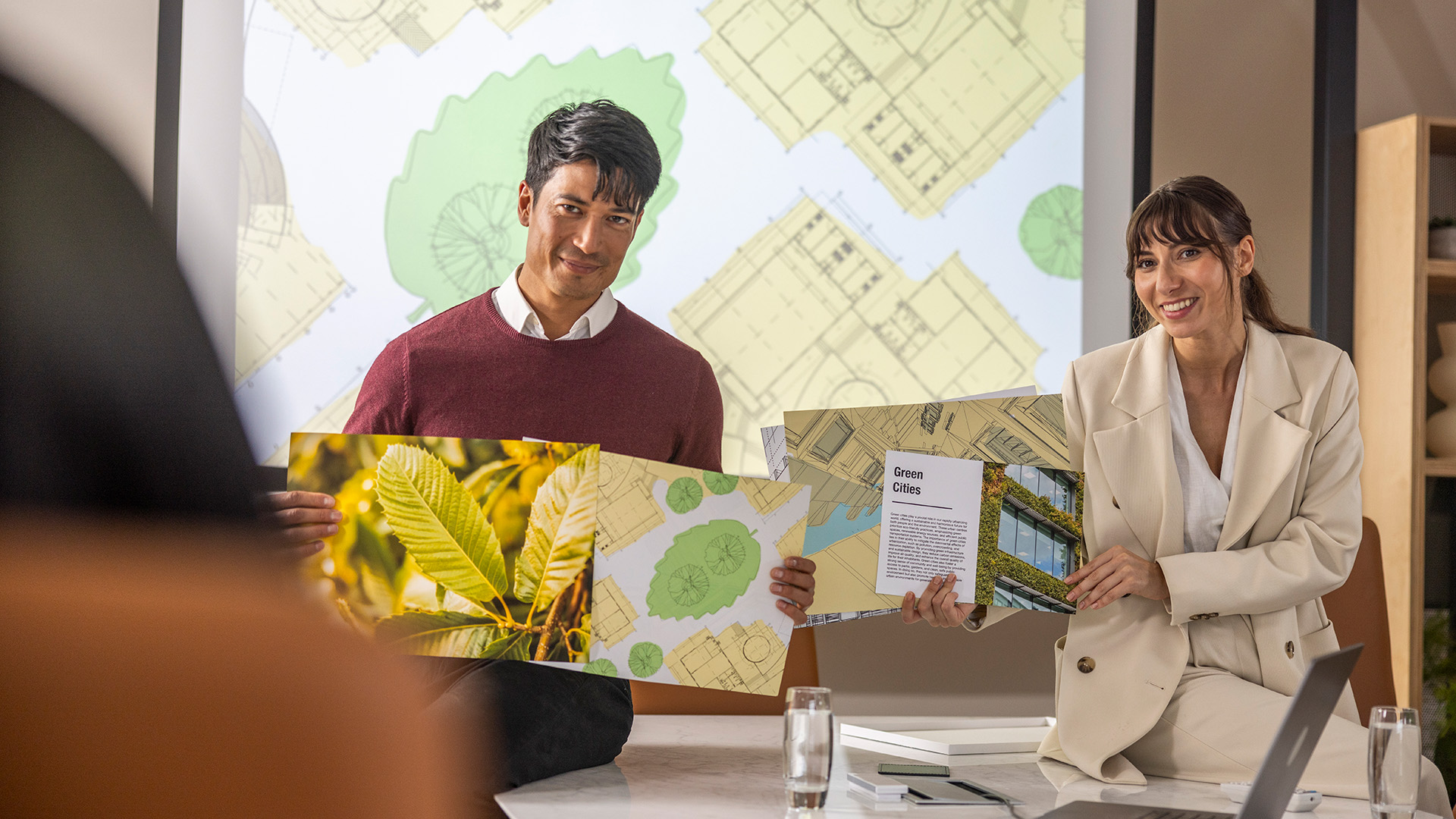 Two people presenting printed sustainability materials in a meeting room, holding large A3 prints featuring nature imagery and architectural plans, with a projected layout displayed behind them.