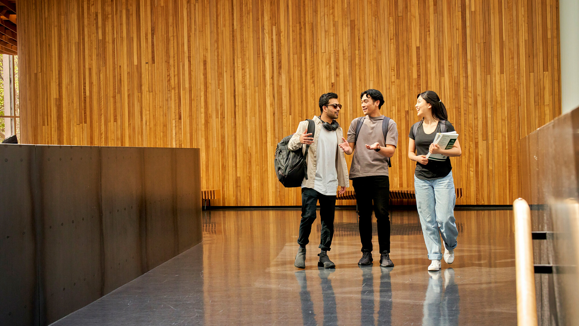 Three tertiary students walking side by side inside a modern building, chatting and carrying books and bags.