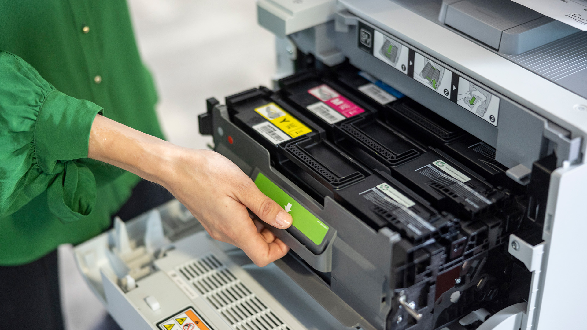 A person removes a toner cartridge from a Brother printer, showing the internal colour cartridges and labelled installation guides.