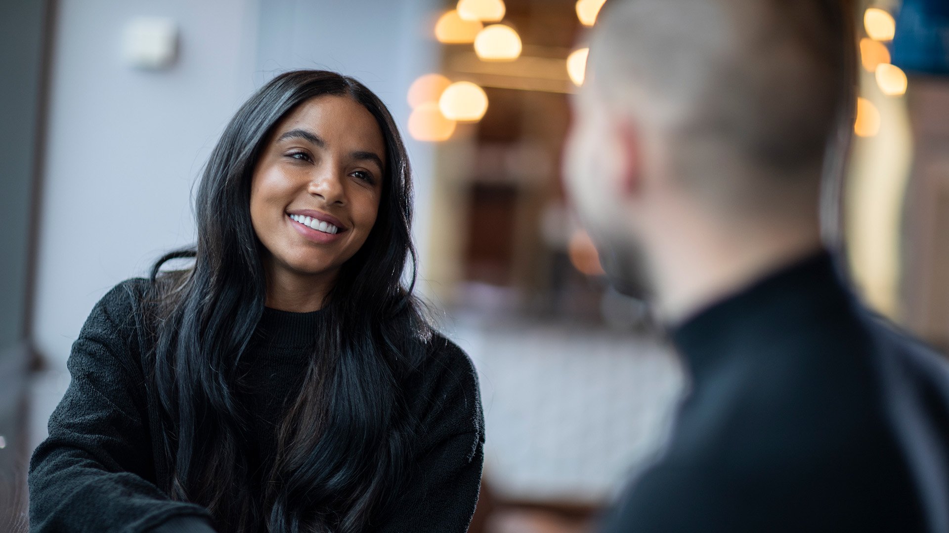 Smiling woman with long dark hair talking to a person in the foreground in a softly lit indoor setting.